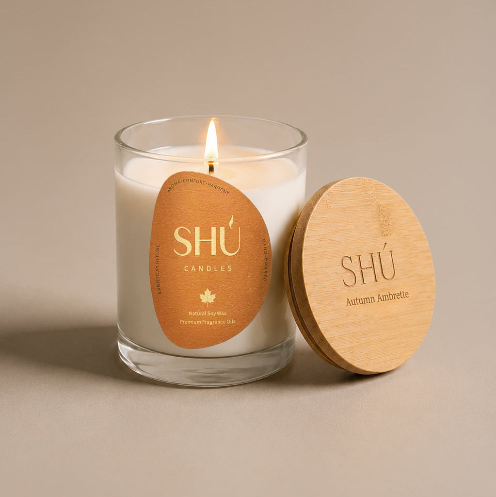 A lit white soy candle in a clear glass jar with a round tan paper label reading 'SHÚ CANDLE ' and 'Pumpkin Noir,' shown beside a light wood lid engraved with the same branding on a warm beige background.