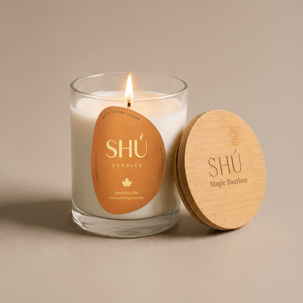 A lit white soy candle in a clear glass jar with a round tan paper label reading 'SHÚ CANDLE ' and 'Pumpkin Noir,' shown beside a light wood lid engraved with the same branding on a warm beige background.