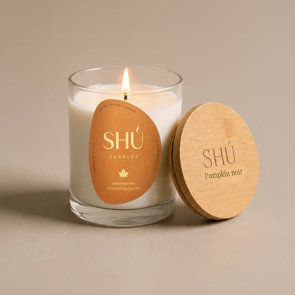 A lit white soy candle in a clear glass jar with a round tan paper label reading 'SHÚ CANDLE ' and 'Pumpkin Noir,' shown beside a light wood lid engraved with the same branding on a warm beige background.